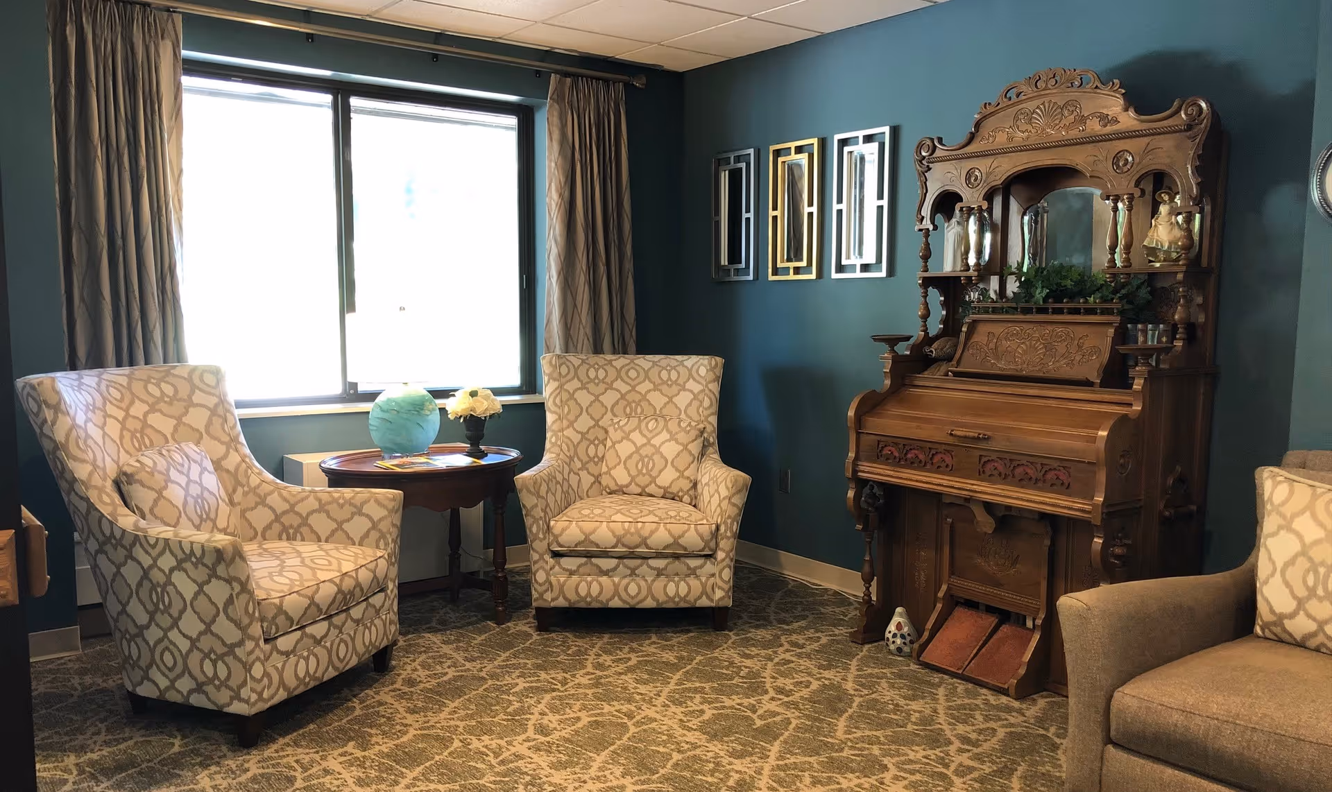 A cozy sitting area in a senior living facility with two patterned armchairs and a small round wooden table between them, placed in front of a window with beige curtains. To the right, there is an ornate wooden antique pump organ with decorative carvings and a few small decorative items on top. The walls are painted teal, and there are three modern rectangular mirrors hanging on the wall above the organ. The floor is carpeted with a patterned design.