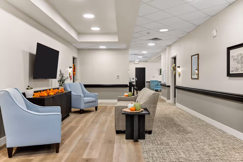 Bright senior living facility lobby with light blue chairs, a beige sofa, a wall-mounted TV, and autumn pumpkins on side tables.