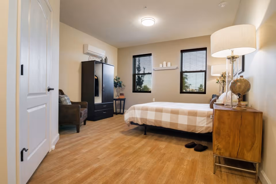 A cozy bedroom with a bed covered in a checkered beige and white blanket, a wooden nightstand with a globe and a lamp, two windows with blinds, a dark wardrobe, a brown armchair with a plaid cushion, and wooden flooring.