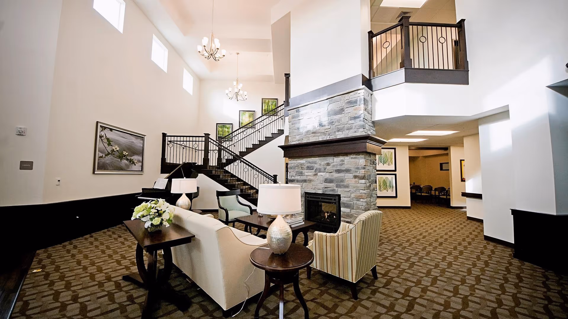 Spacious assisted-living lobby with seating arranged around a stone fireplace, a staircase, and chandeliers.