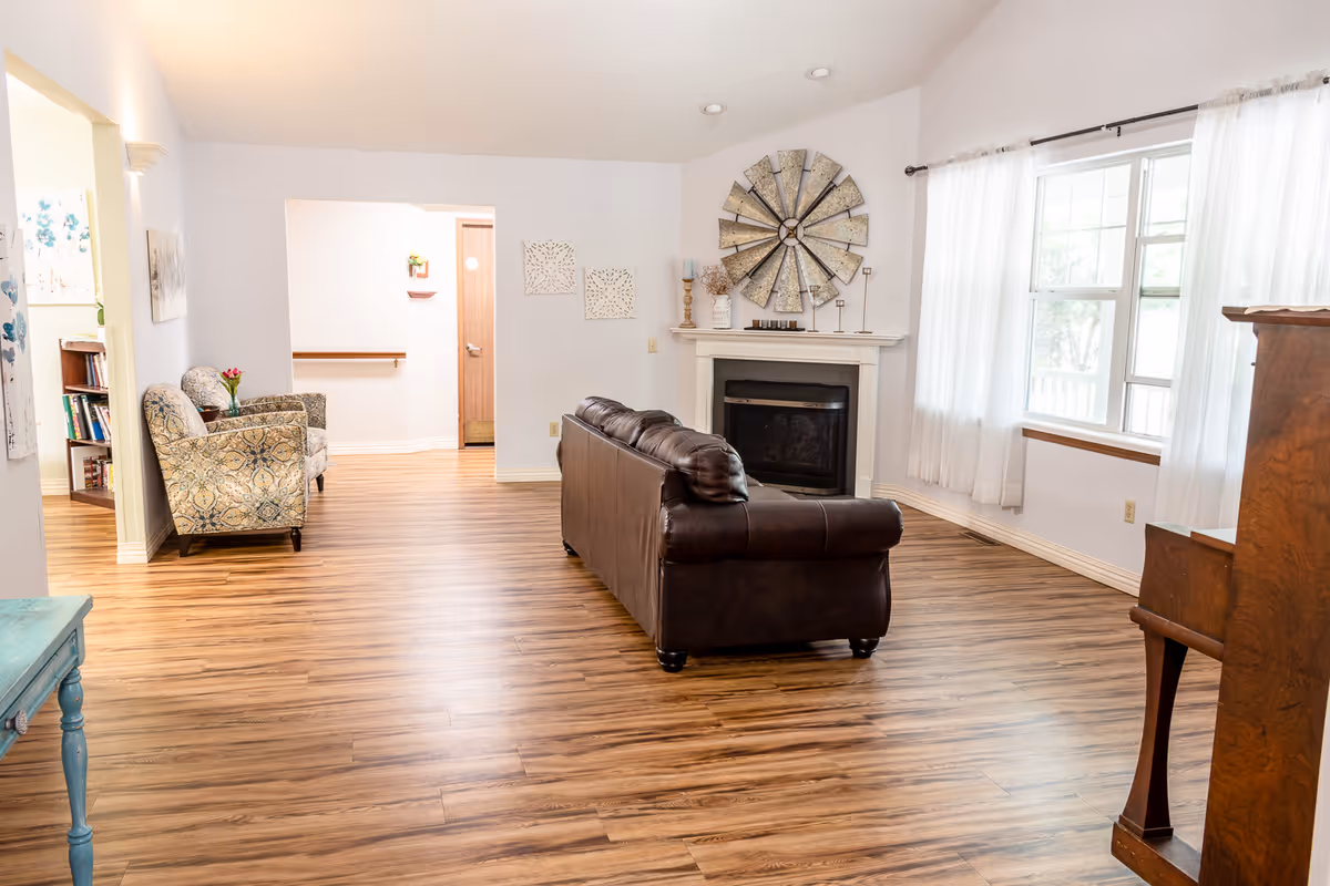 Bright open living room with a leather sofa facing a fireplace, patterned armchairs, a large window with sheer curtains, and wood-look flooring.