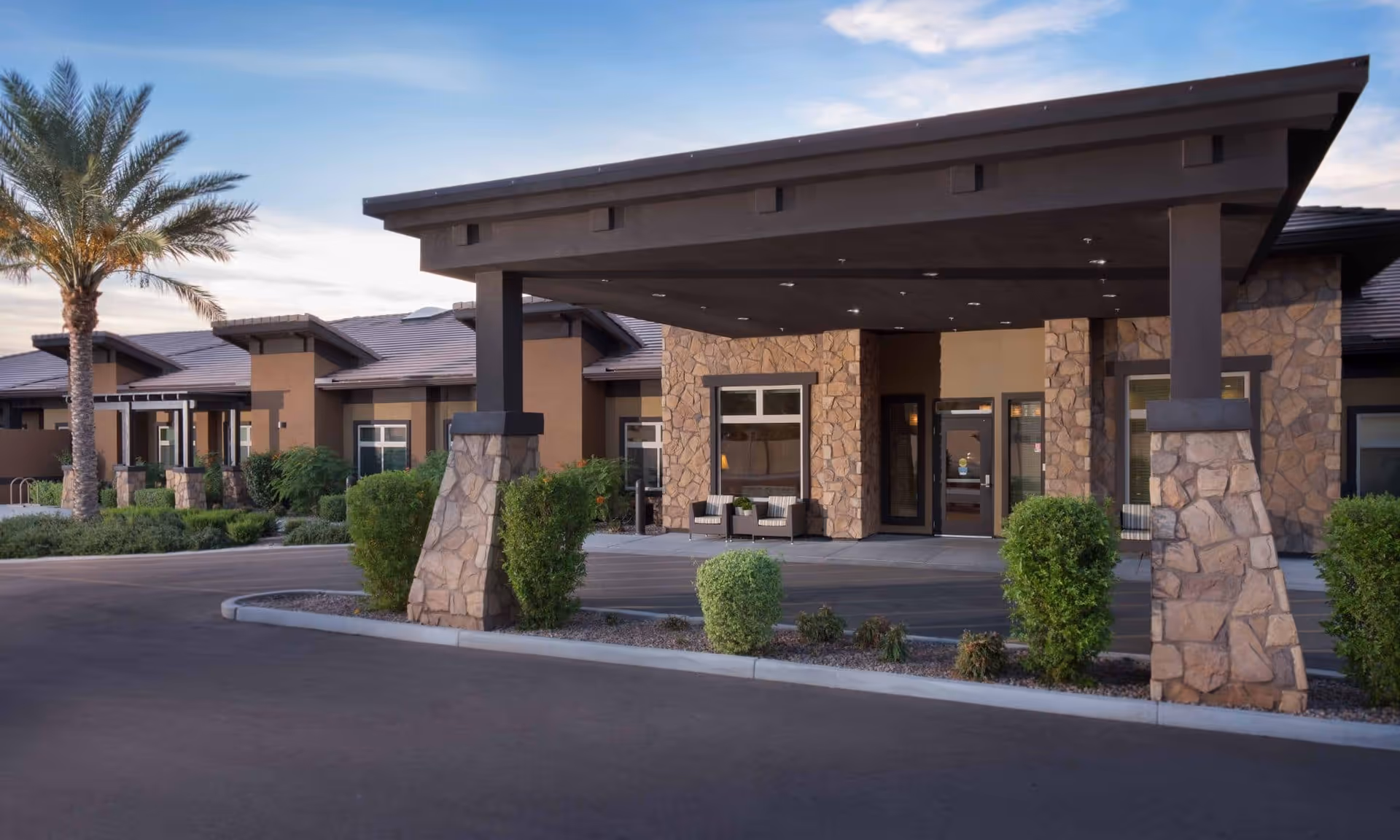 Exterior view of Chandler Memory Care facility entrance with a covered drop-off area supported by stone pillars, surrounded by landscaped bushes and a palm tree under a clear sky.