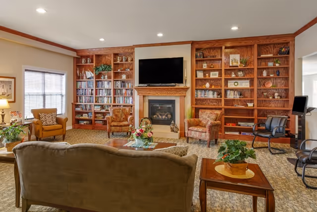 A cozy living room with a large wooden bookshelf filled with books and decorative items, a fireplace with a TV mounted above it, several upholstered armchairs, a sofa, and a wooden coffee table with a floral centerpiece. The room has carpeted flooring and recessed ceiling lights.