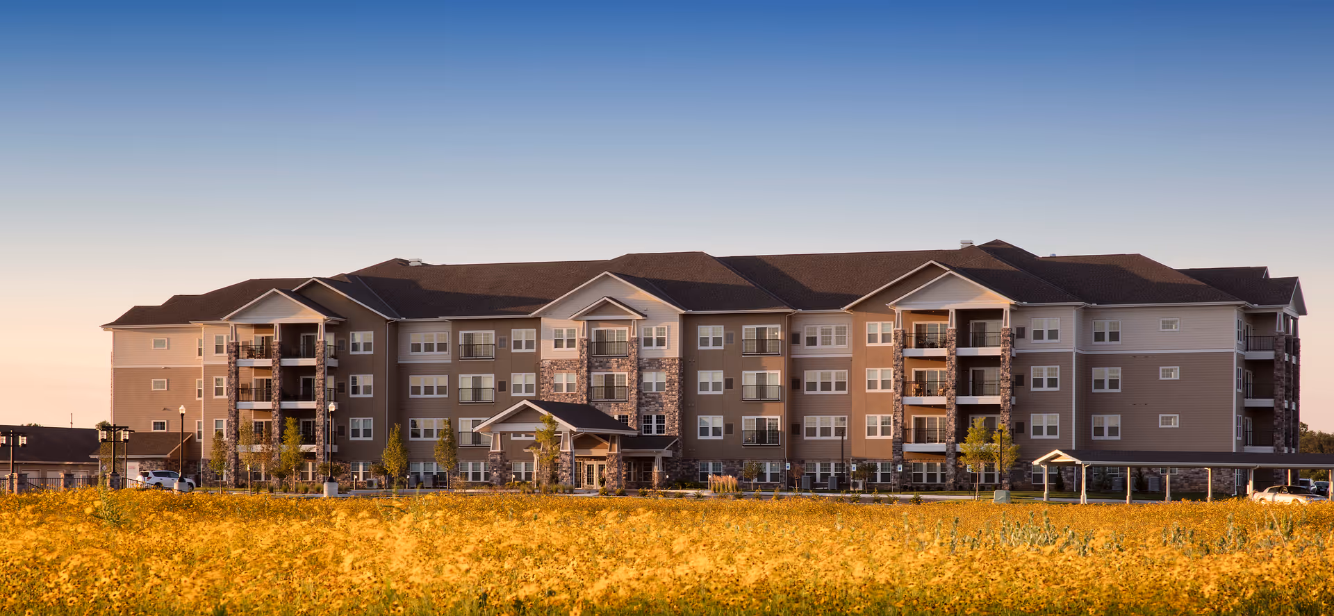 A large, multi-story senior living apartment building with balconies, set behind a field of yellow flowers under a clear blue sky.