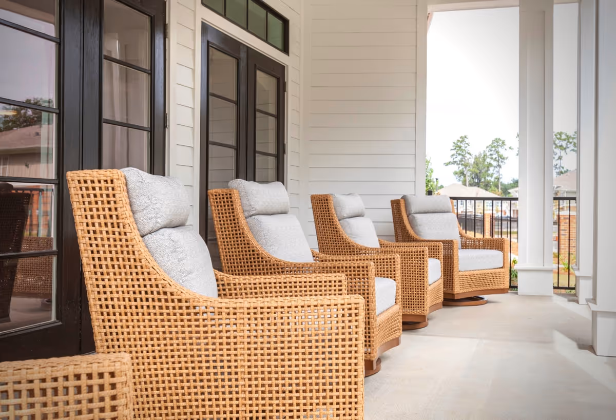A row of four wicker armchairs with light gray cushions arranged on a covered porch with white walls and black-framed glass doors, overlooking a fenced outdoor area with trees and buildings in the background.