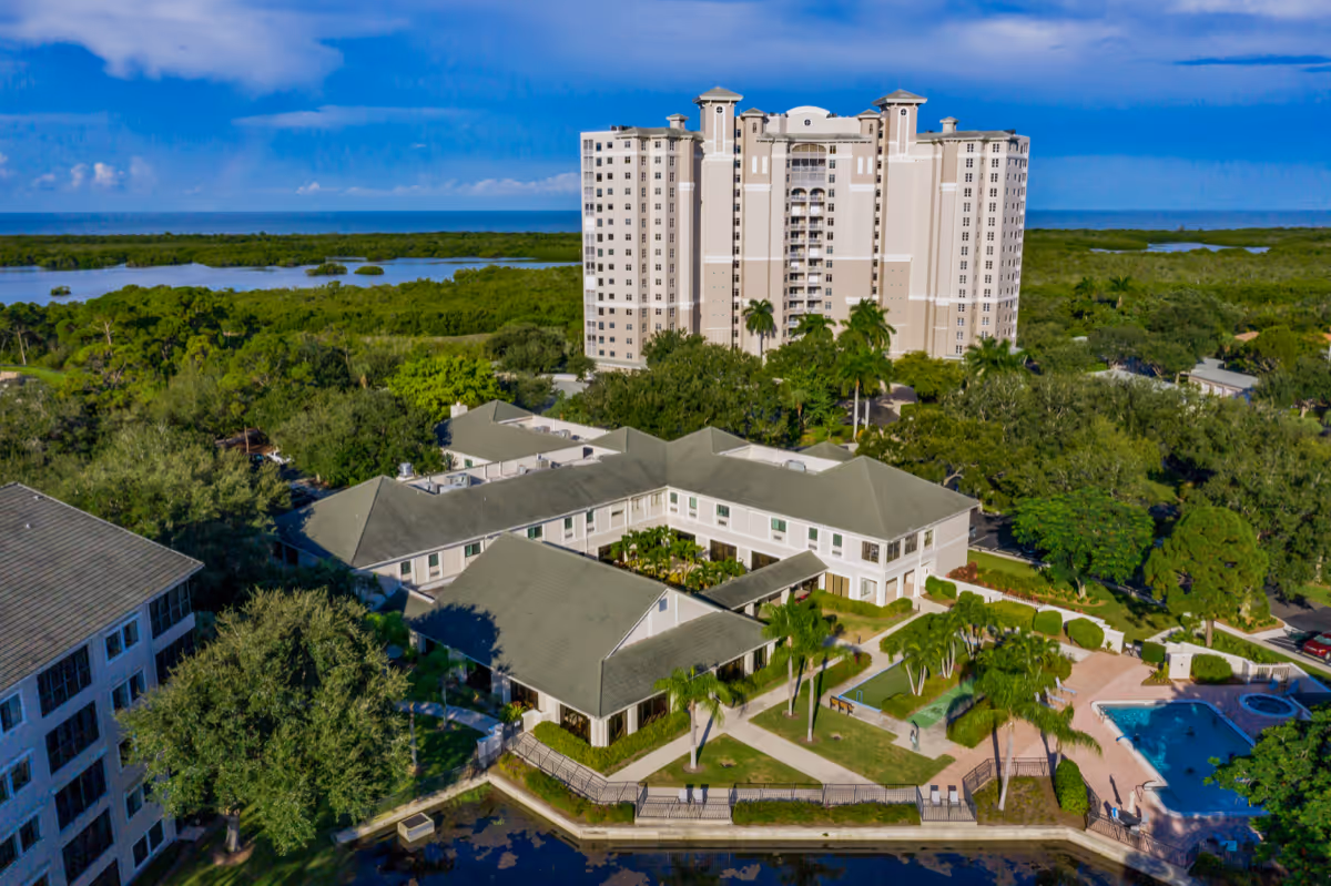 Aerial view of a senior living campus with low-rise buildings, a tall tower, landscaped grounds, a swimming pool, and a waterway.