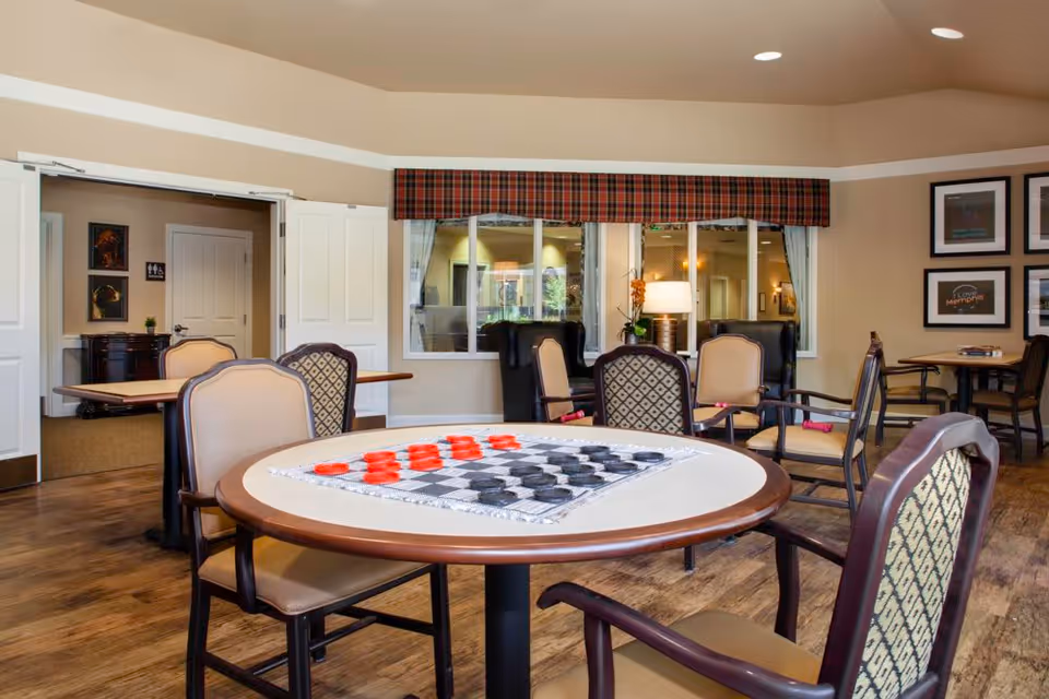 A cozy common area in a memory care community featuring a round table with a checkers game set up, surrounded by cushioned chairs. The room has wood flooring, beige walls with white trim, and a window with a plaid valance. Additional seating and tables are visible in the background, along with framed artwork on the walls and a lamp on a side table.