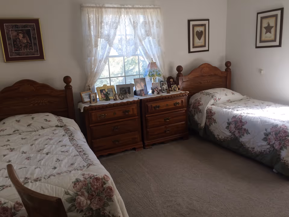 Two twin beds with floral bedspreads and wooden headboards flanking wooden dressers topped with framed photos beneath a curtained window.