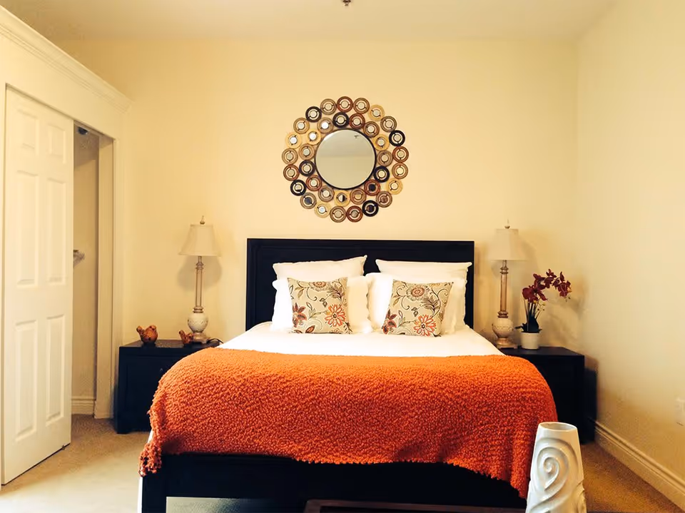 A neatly made bed with white pillows and floral decorative cushions, covered with an orange blanket. On either side of the bed are dark wooden nightstands with matching lamps. Above the bed is a circular decorative mirror with a multi-colored frame. The room has light-colored walls and carpet, with a partially open white door on the left and a white vase with a spiral design in the foreground on the right.