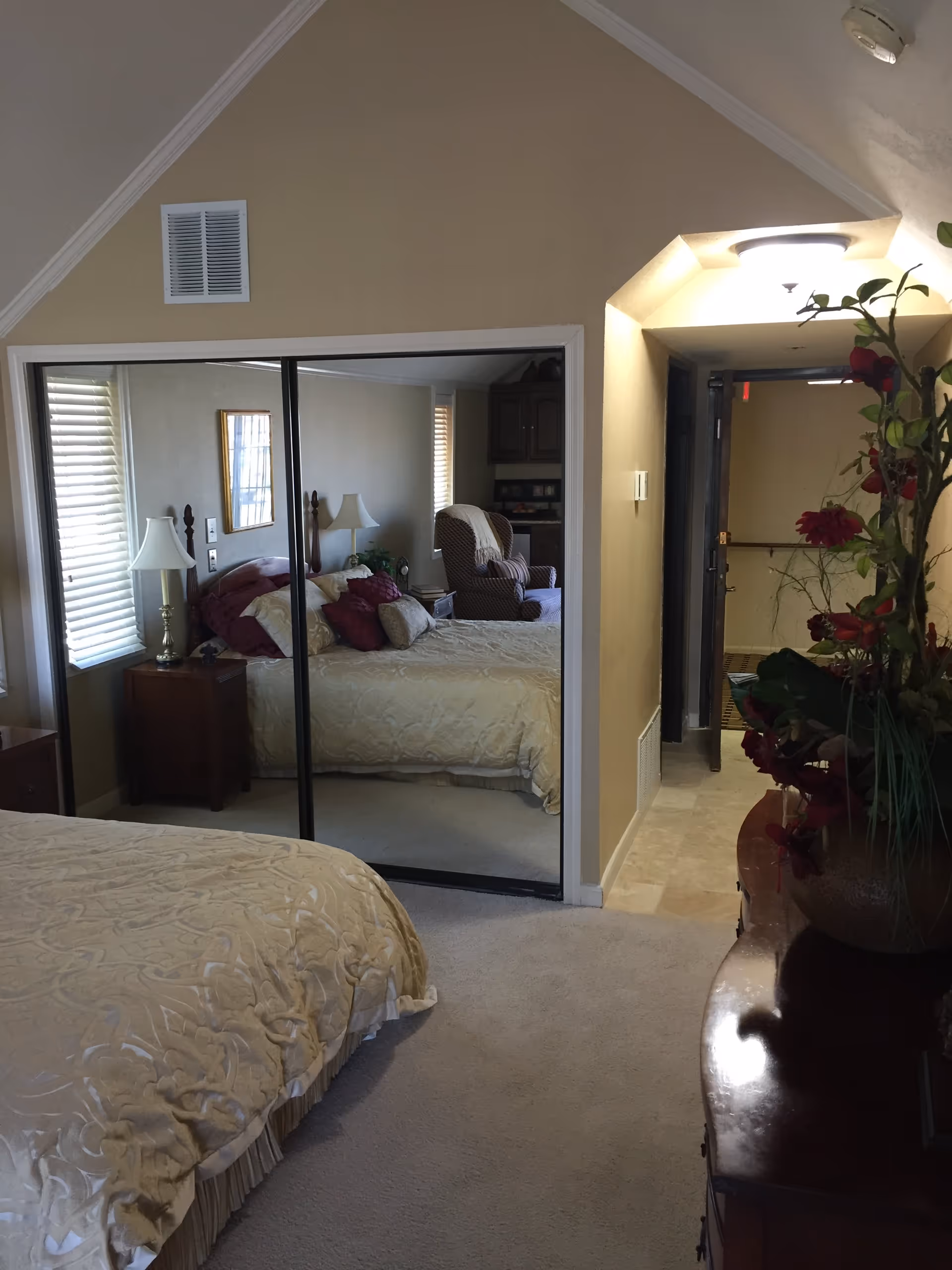 A bedroom with a beige bedspread on the bed, a wooden nightstand with a lamp, and a mirrored closet door reflecting the bed and a cushioned armchair. There is a dresser with a vase of red flowers on the right side and a hallway leading to another room with a ceiling light above the entrance.