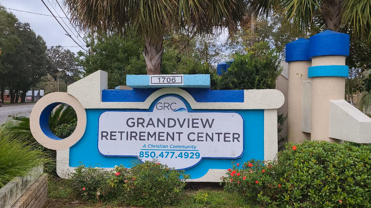 Outdoor sign for Grandview Retirement Center, a Christian community, with the phone number 850-477-4929. The sign is surrounded by greenery and flowers, with a palm tree and other trees in the background.