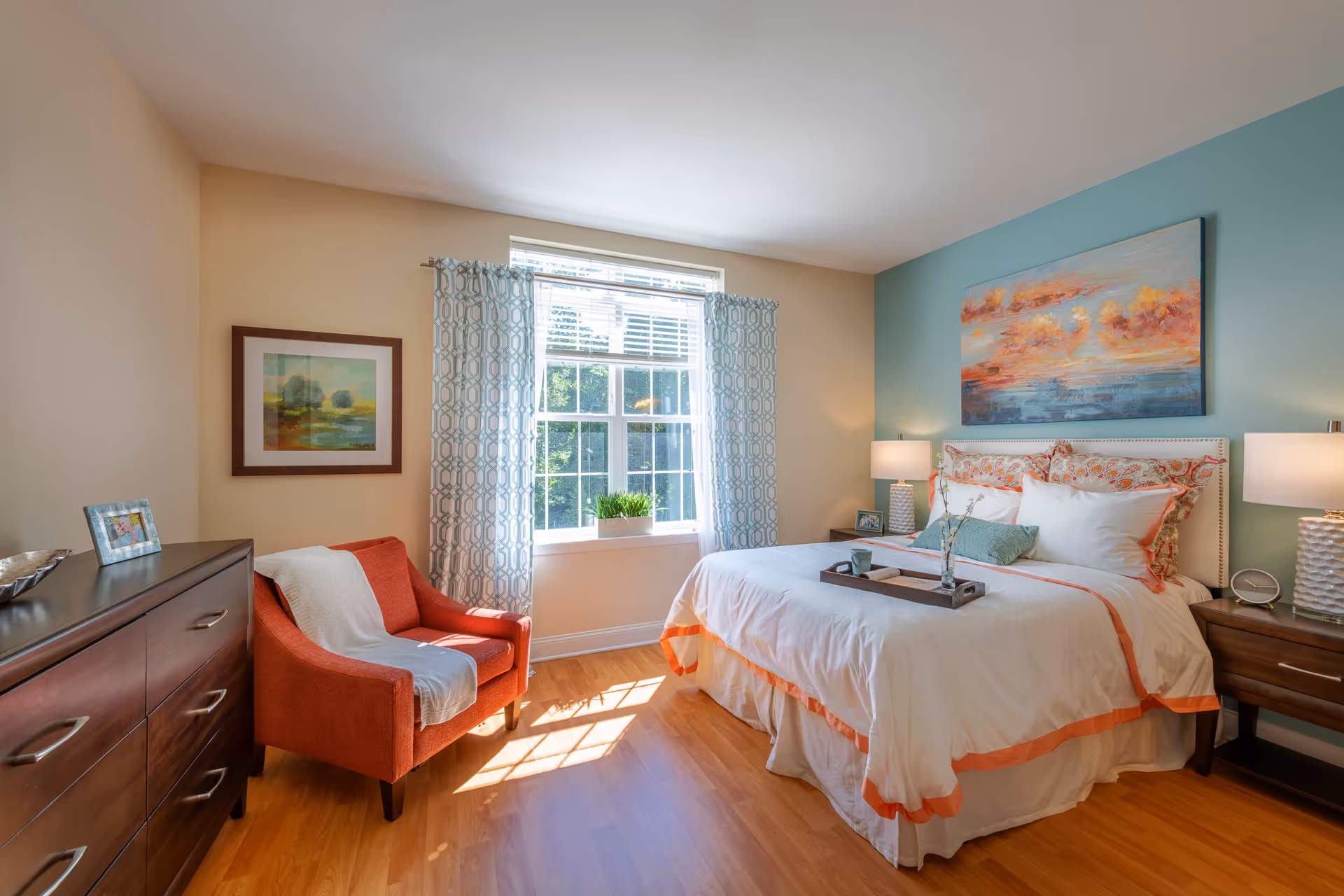 Sunlit bedroom with a neatly made bed, nightstands and lamps, an orange armchair, dresser, and a window with patterned curtains.