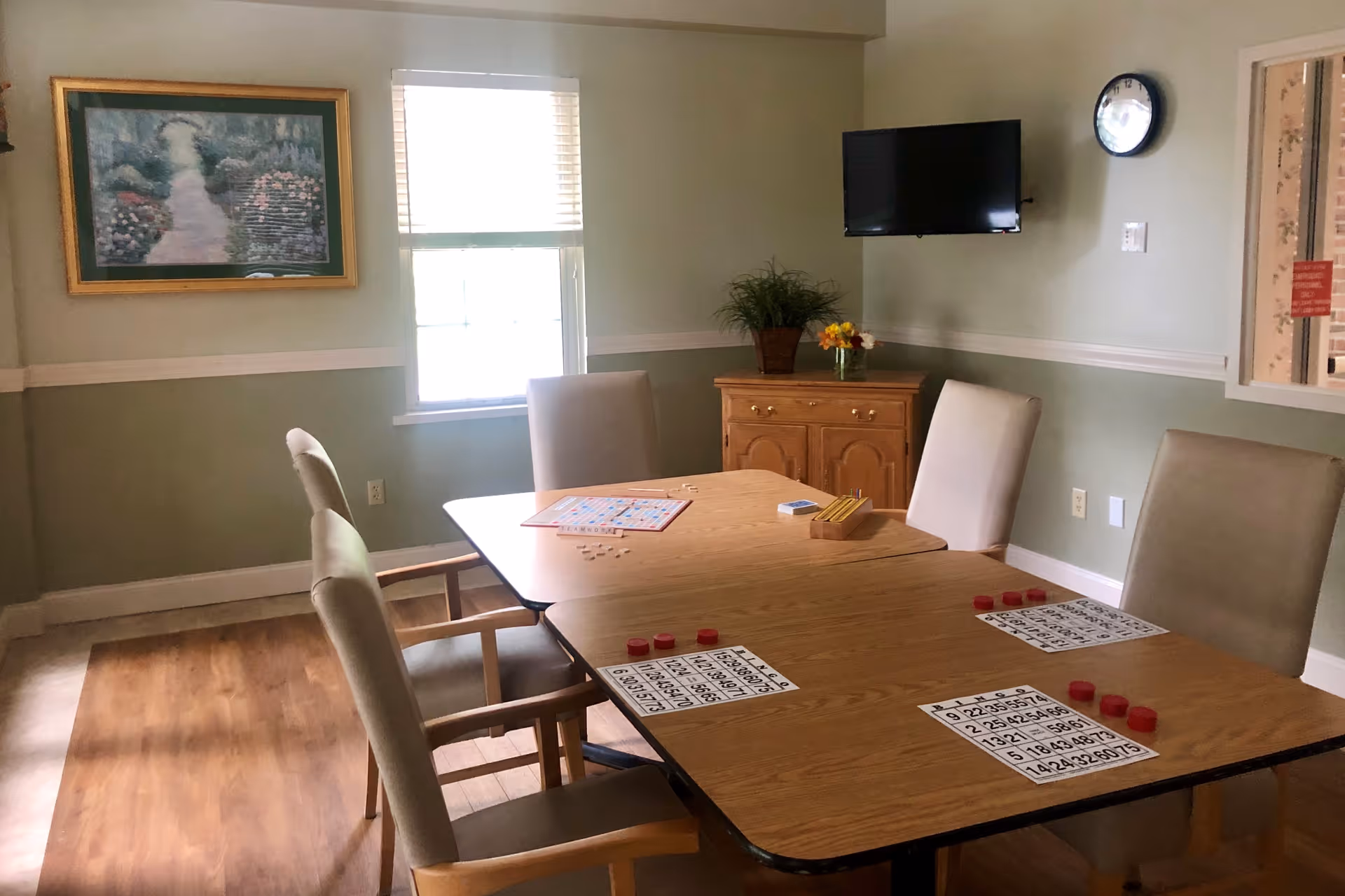 A well-lit room with a wooden table set up for a bingo game, featuring bingo cards and red markers. Four beige upholstered chairs surround the table. The room has light green walls with white trim, a window with blinds, a wall-mounted TV, a clock, a wooden cabinet with a potted plant and a small flower arrangement, and a framed painting on the wall.