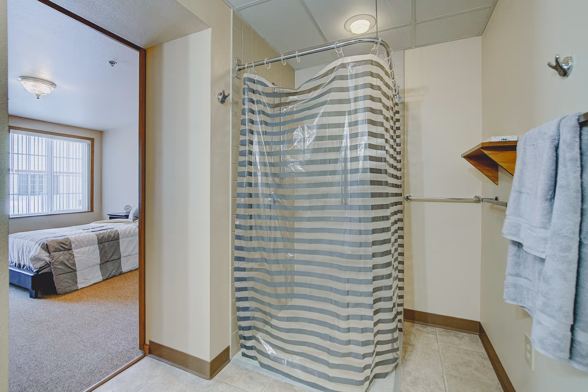 A bathroom with a shower enclosed by a striped shower curtain. There are two gray towels hanging on a towel rack on the right wall, and a small wooden shelf above the towels. The bathroom floor is tiled. Through the open doorway, a bedroom with a bed covered in a gray and white checkered blanket is visible, along with a window with vertical blinds.