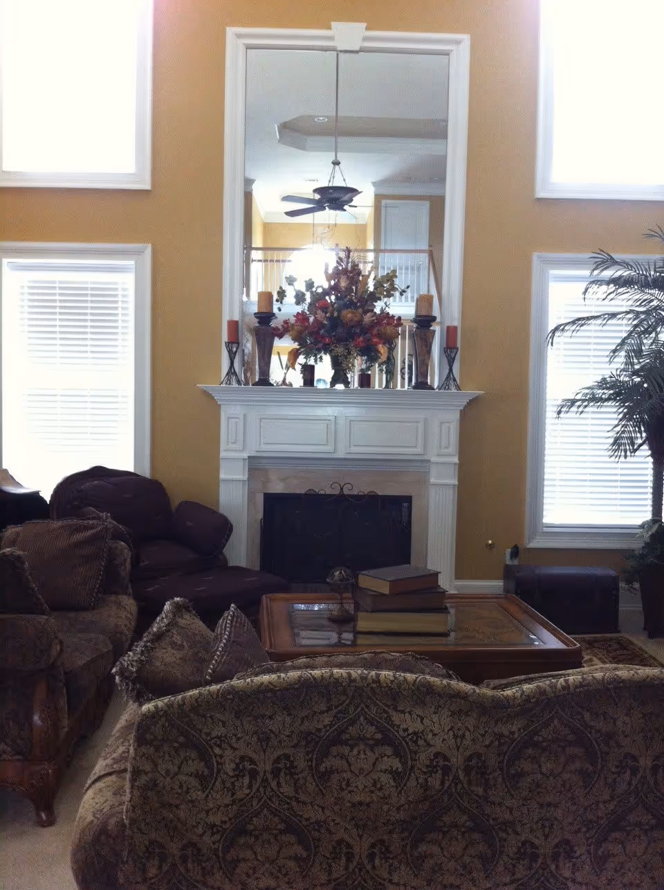 A cozy living room with patterned upholstered sofas and armchairs arranged around a wooden coffee table with stacked books. A white fireplace with a decorative screen is centered on the far wall, topped with a large mirror and a floral arrangement flanked by candles. The room has tall windows with white blinds and a potted plant on the right side.