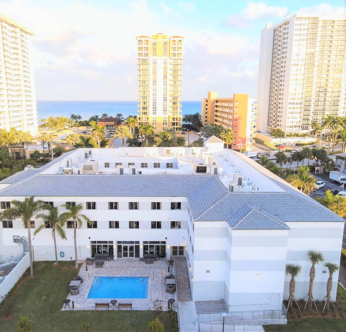 Aerial view of a white senior living building with a central pool courtyard, palm trees, and ocean-front high-rises in the background.