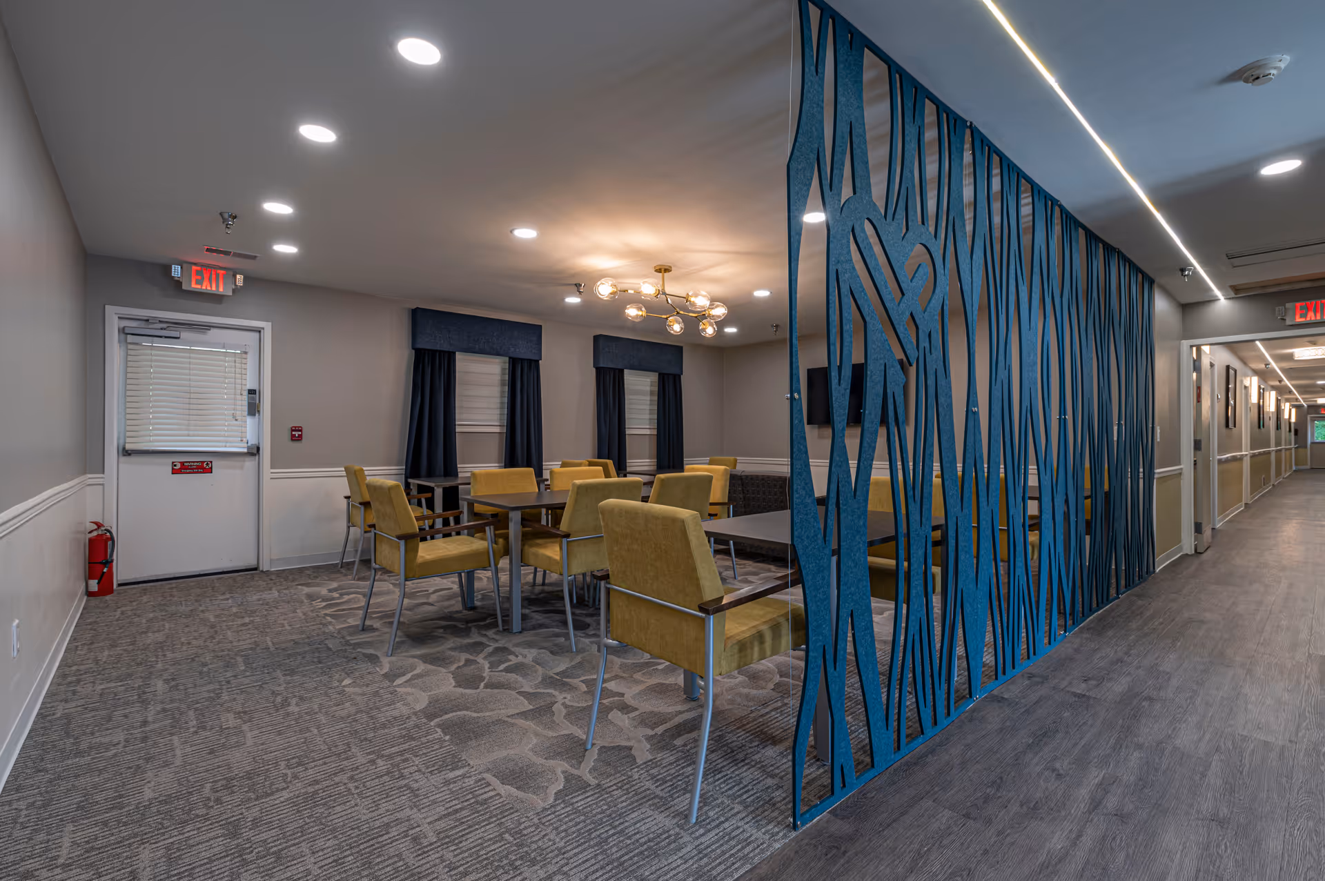 Interior view of a senior living facility dining area with yellow cushioned chairs around tables, a decorative blue partition, carpeted and wood flooring, ceiling lights, and an exit door in the background.