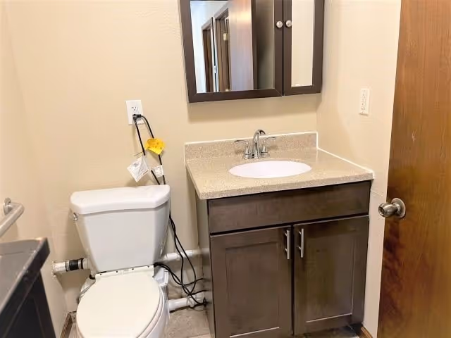 Small bathroom with a toilet, vanity with sink and mirrored medicine cabinet, and exposed plumbing and cords.