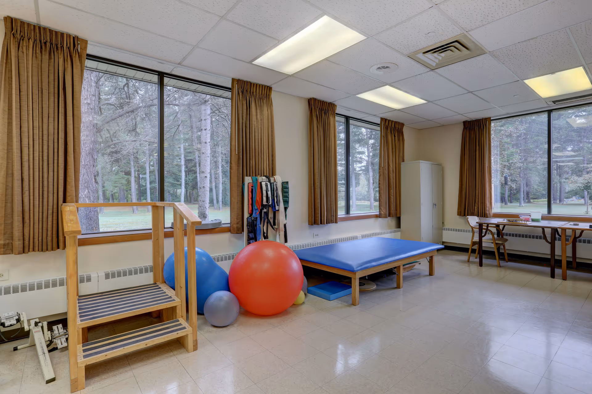A physical therapy room with large windows showing a wooded outdoor area. The room contains a wooden step platform, several exercise balls in different sizes and colors, a padded therapy table, and a small table with chairs near the windows. Brown curtains hang beside the windows.