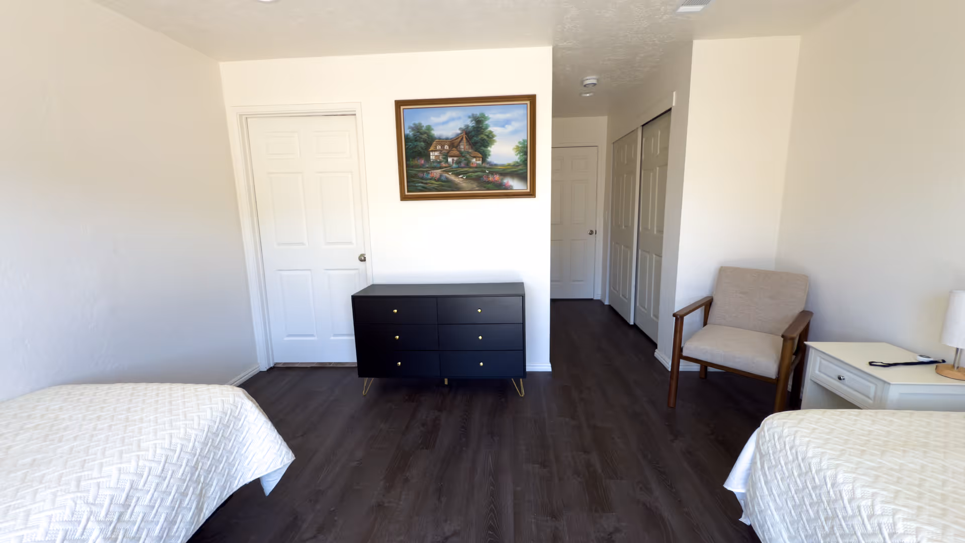A clean and simple bedroom with two twin beds covered in white textured bedspreads. The room has dark wood flooring, a black dresser with six drawers against the far wall, a beige armchair next to a white nightstand with a lamp and a remote control on it. There is a framed painting of a house and garden hanging above the dresser. Several white doors are visible, including a closet with double doors and a door leading out of the room.