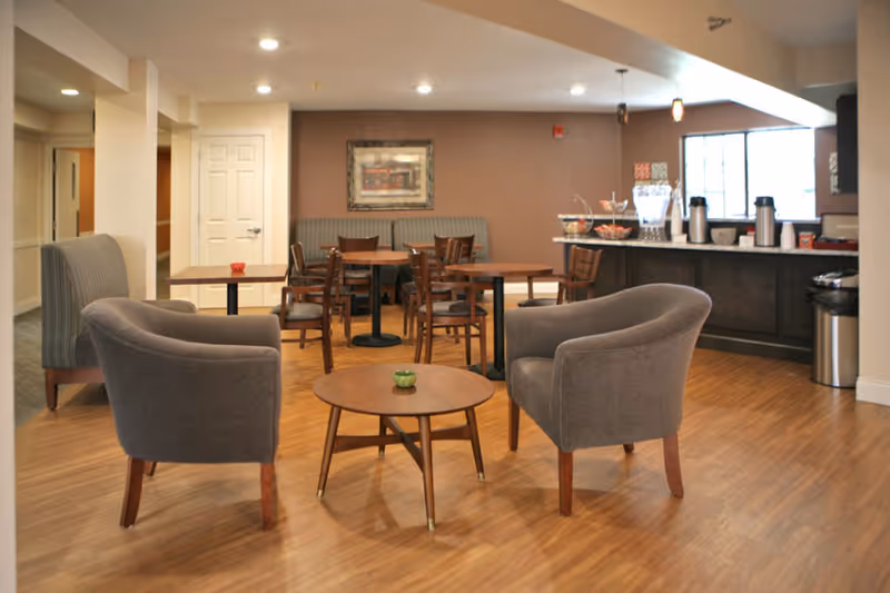 A furnished senior living common area with two gray armchairs around a round coffee table and dining tables and a service counter in the background.