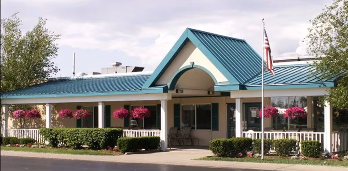 Front exterior view of Eden Heights of West Seneca Assisted Living building with a teal metal roof, white pillars, hanging flower baskets with pink flowers, trimmed bushes, and an American flag on a flagpole in front.