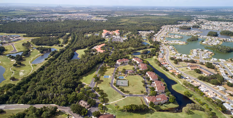 Aerial view of a senior living facility named Freedom Plaza surrounded by greenery, water bodies, and residential areas. The facility buildings are arranged around open green spaces with tennis courts and walking paths. Nearby are wooded areas, a golf course, and a suburban neighborhood with houses and roads.