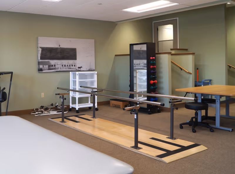 Physical therapy room with parallel walking bars on a wooden platform, a rack of dumbbells, a treatment table, and a desk.
