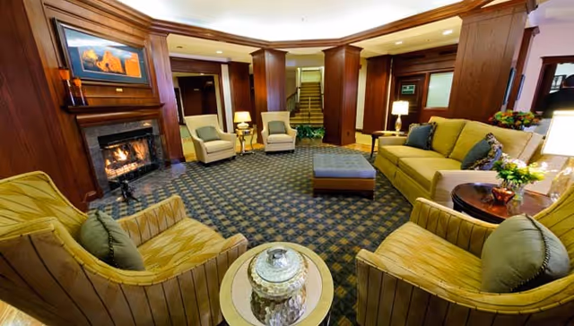 A cozy living room area with a lit fireplace, wooden paneling, and a patterned carpet. The room features a green sofa, two armchairs with green cushions, two beige armchairs, a blue ottoman, side tables with lamps, and a decorative jar on a round table. There are stairs and doorways in the background.