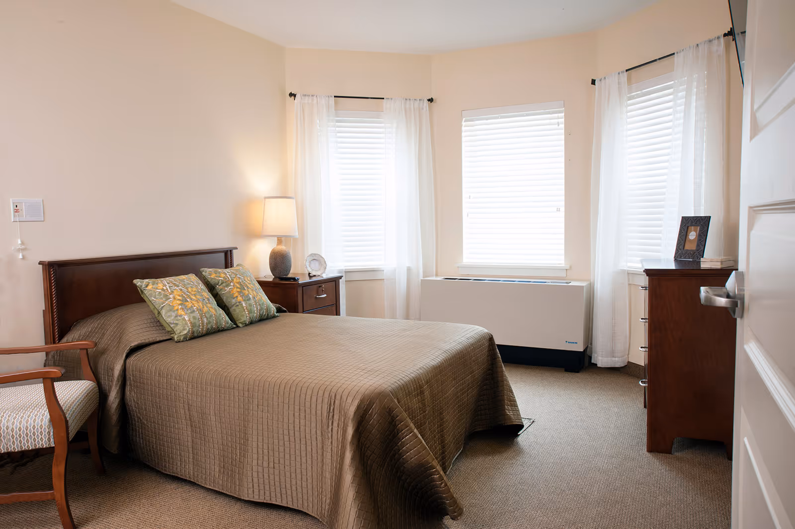 Sunlit bedroom with a neatly made bed, bedside table and lamp, dresser, chair, and three windows with blinds and sheer curtains.