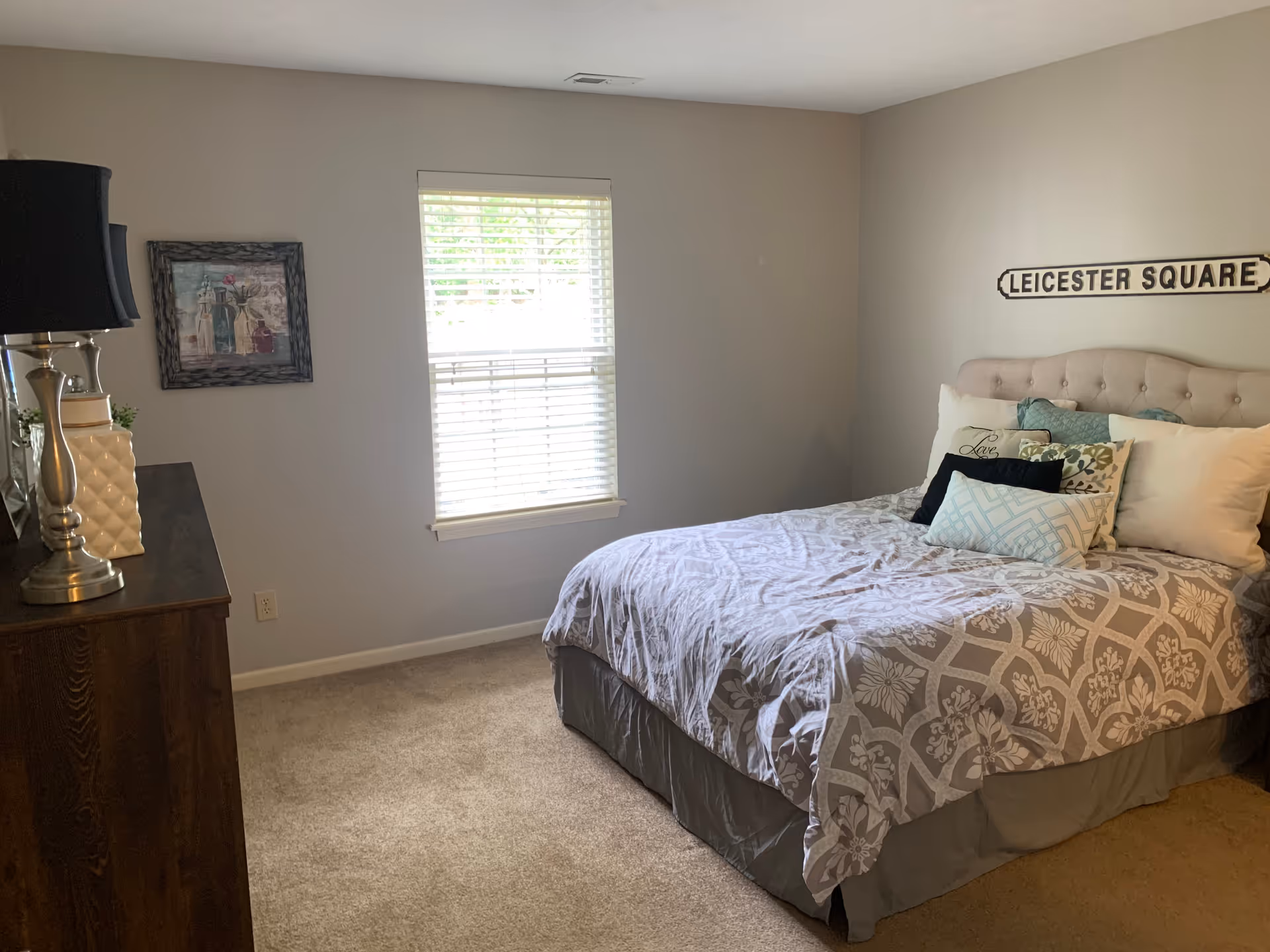 A bedroom with a bed covered in a patterned gray and white comforter and multiple pillows. Above the bed is a wall decoration that reads 'LEICESTER SQUARE'. To the left is a wooden dresser with a silver lamp and decorative items. A window with white blinds is centered on the back wall, and a framed picture hangs to the left of the window. The room has beige carpet and light gray walls.