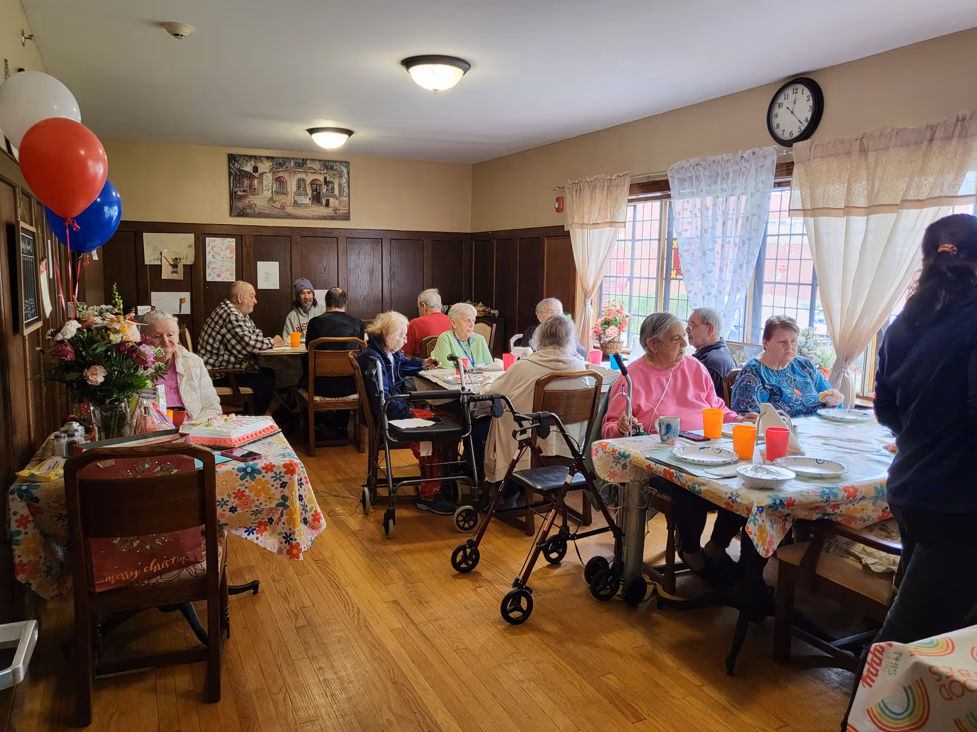 A group of elderly people sitting around tables covered with floral tablecloths in a well-lit room with wooden paneling on the walls. Some individuals are using walkers and wheelchairs. There are colorful balloons and flowers on one table, and a clock on the wall above windows with sheer curtains letting in natural light.
