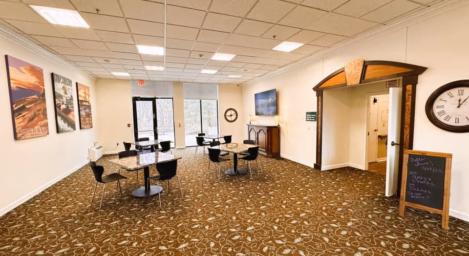 A spacious room with patterned carpet, several small square tables each surrounded by black chairs, large windows and a glass door letting in natural light. The walls are decorated with framed posters and a clock. There is a wooden cabinet with a TV mounted above it and a chalkboard sign near an arched doorway.