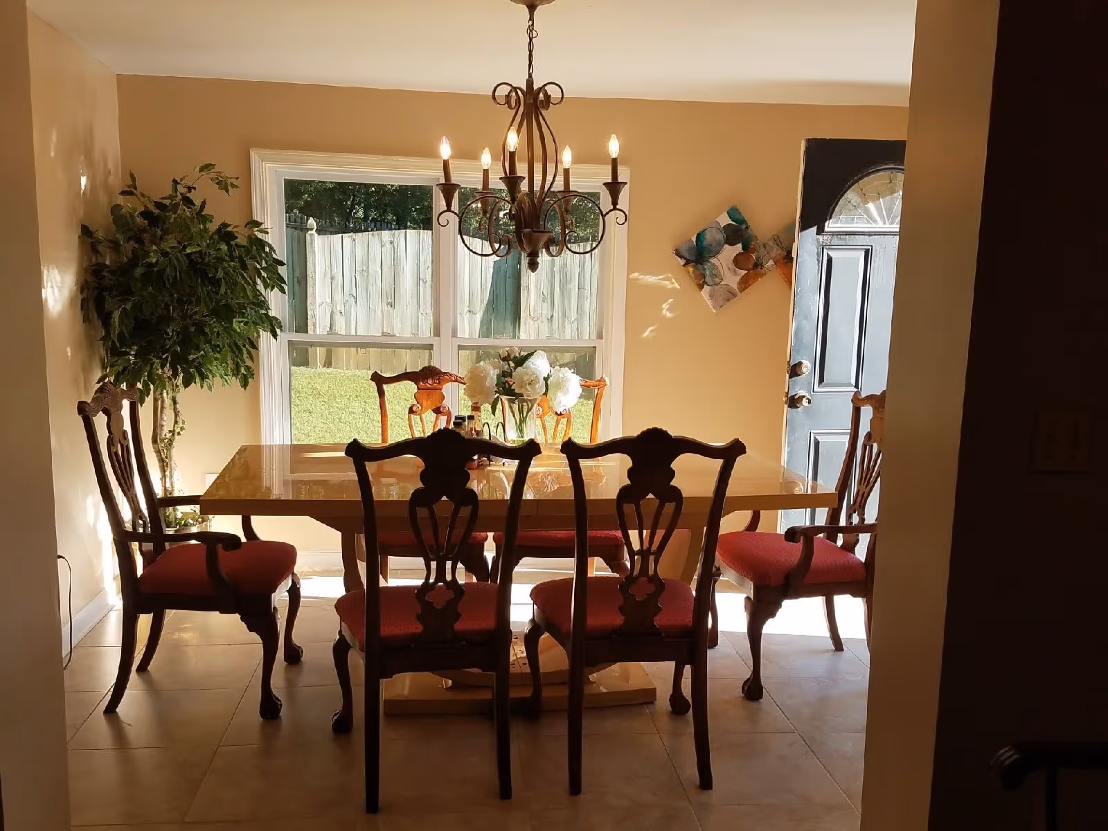 A dining room with a wooden dining table and six chairs with red cushions. A chandelier with lit bulbs hangs above the table. There is a large window behind the table showing a fenced backyard with grass. A potted plant is in the corner to the left, and a partially open black door is on the right wall with a decorative wall art piece nearby.