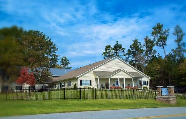 Exterior view of a single-story senior living community building with beige siding, a gabled roof, and a covered front porch. The building is surrounded by a black metal fence and landscaped with bushes and trees. The sky is partly cloudy with blue patches.