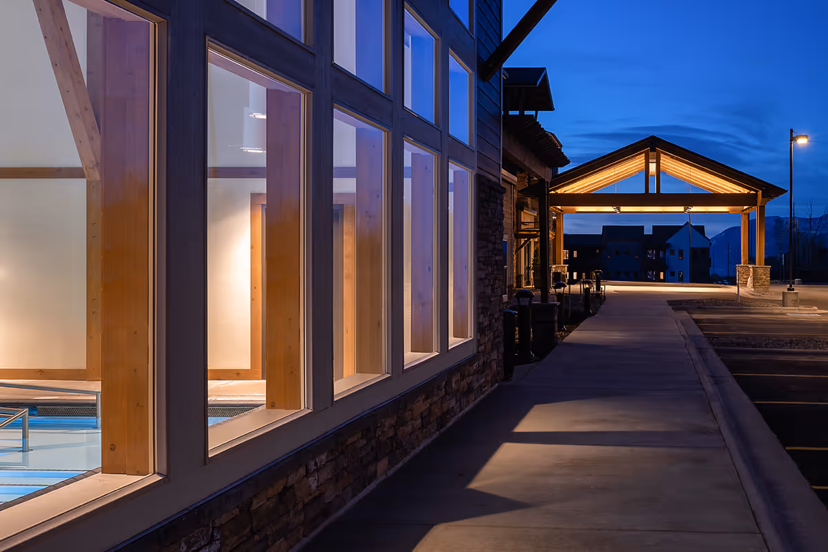 Evening view of the exterior of a building with large windows showing an indoor pool area. The building has wooden beams and stone accents, with a covered entrance illuminated by warm lights. The sky is dark blue, indicating dusk or early night.