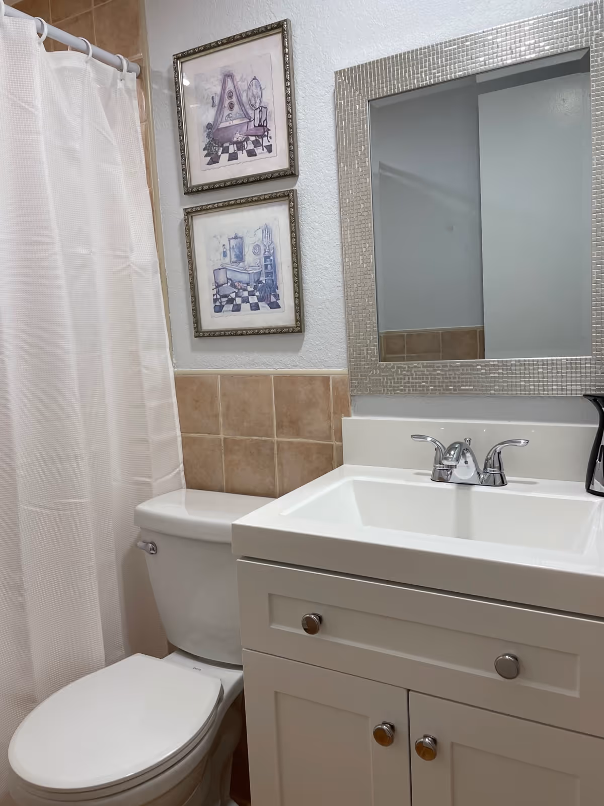 A bathroom with a white toilet and a white vanity with a sink and chrome faucet. Above the vanity is a rectangular mirror with a textured silver frame. To the left of the toilet is a shower with a white curtain. The wall behind the toilet and vanity is partially tiled with beige tiles, and two framed pictures with purple artwork hang above the toilet.