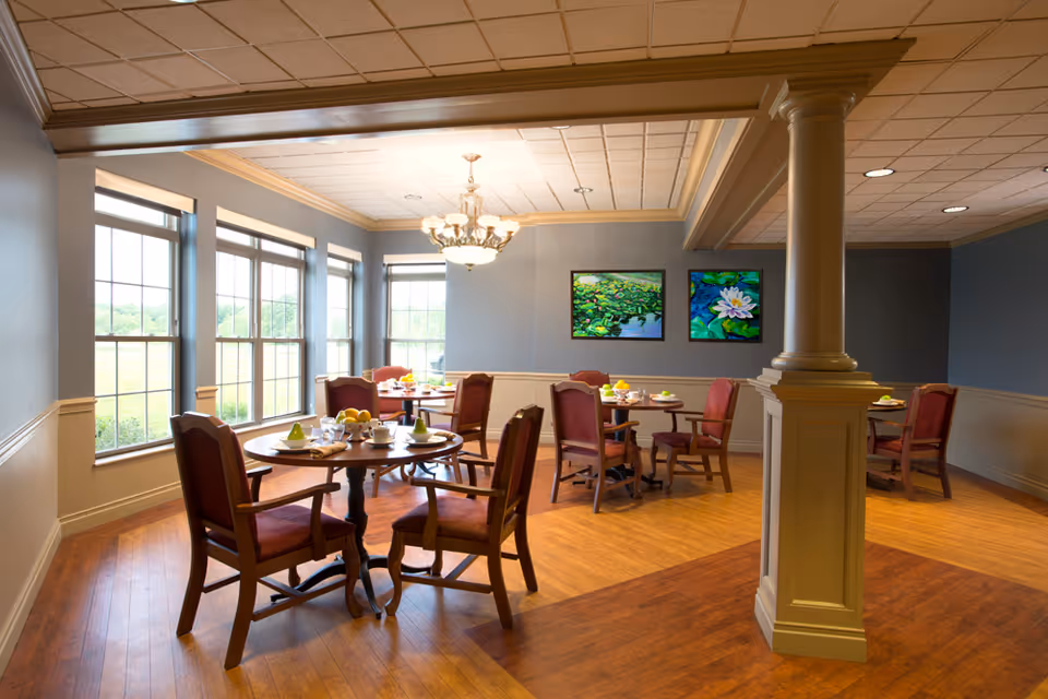 A dining room with several round wooden tables, each set with cups, plates, and fruit. The room has large windows letting in natural light, blue walls with white wainscoting, wooden flooring, a chandelier, and two paintings of water lilies on the wall.