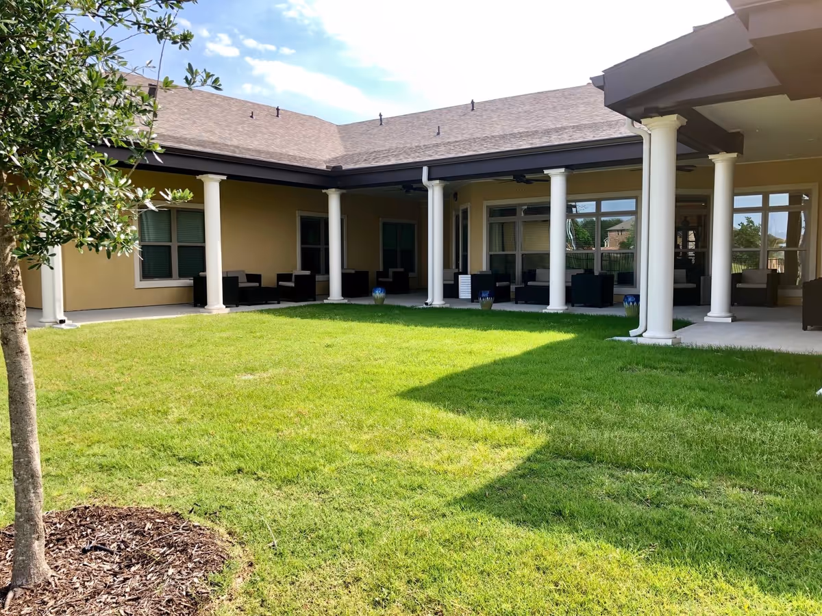 A spacious outdoor courtyard area with green grass, a small tree on the left, and a covered patio supported by white columns. The patio has several black cushioned chairs and small tables arranged along the walls of a beige building with multiple windows.
