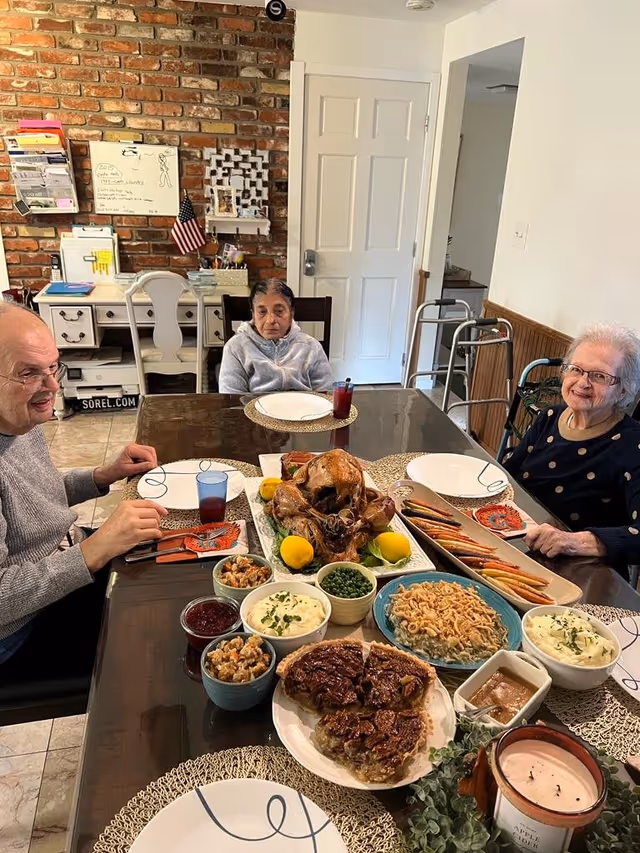 Three elderly people sitting around a dining table set with a Thanksgiving meal including a roasted turkey, pecan pie, mashed potatoes, peas, cranberry sauce, and other side dishes in a cozy dining room with a brick wall and white door in the background.