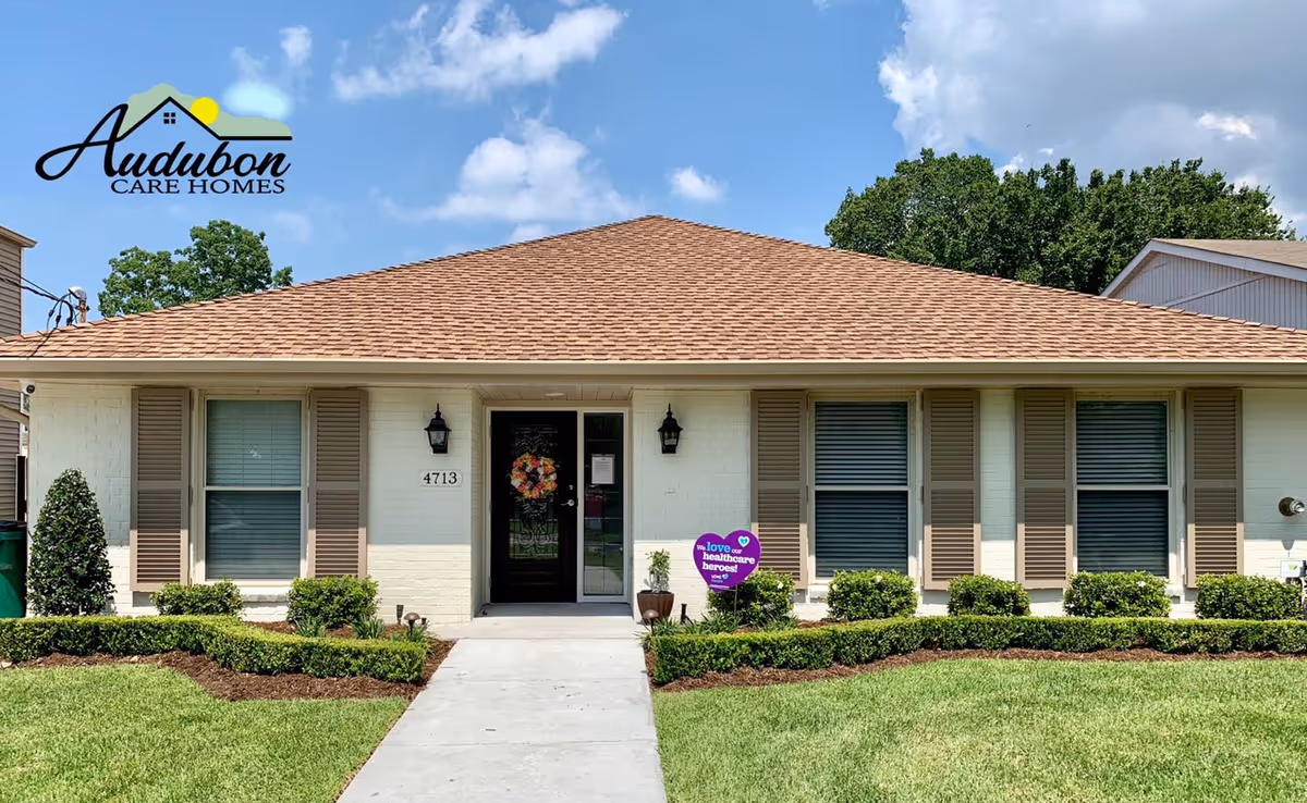 Single-story brick residential building with a central glass door, shuttered windows, trimmed hedges and a sidewalk leading to the entrance.