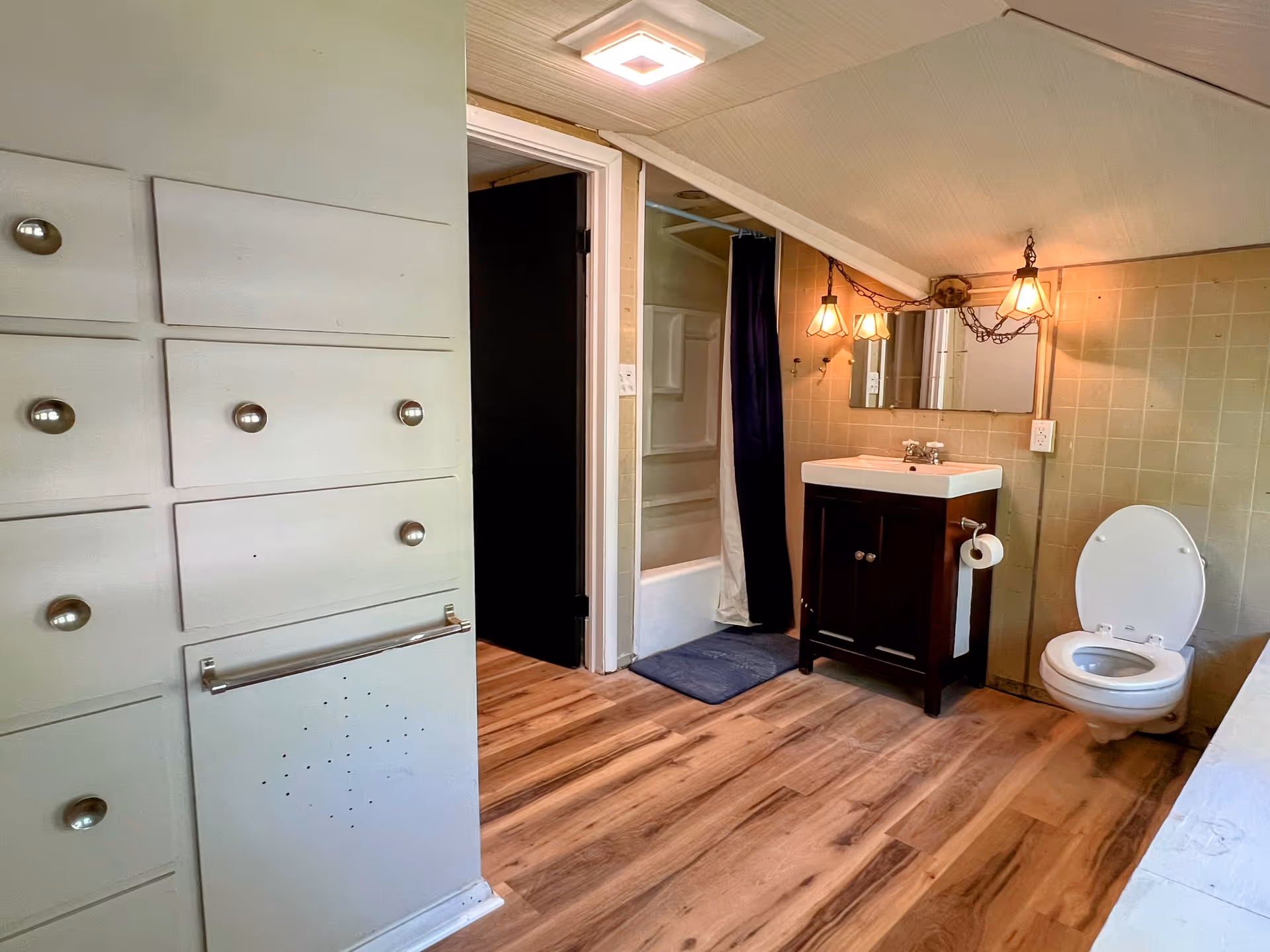 A bathroom with wooden flooring, a white toilet with the lid open, a dark wood vanity with a white sink and a mirror above it, two hanging light fixtures, a bathtub with a shower curtain, and built-in cabinets with silver knobs on the left side.