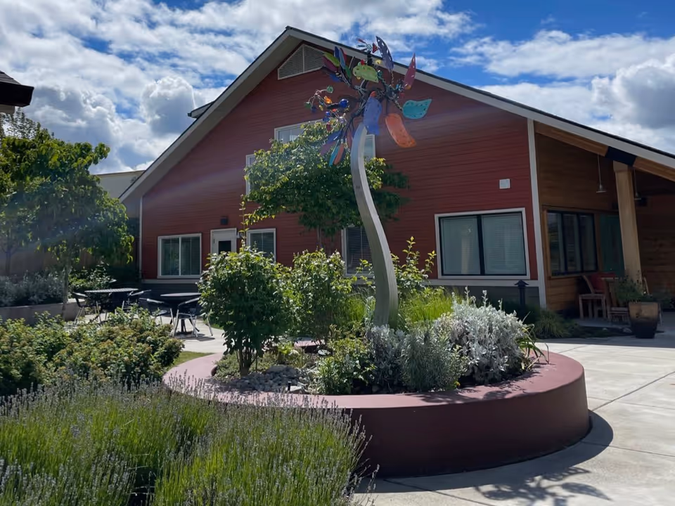 Outdoor courtyard with a circular planter and colorful sculpture in front of a red building with seating under a partly cloudy sky.