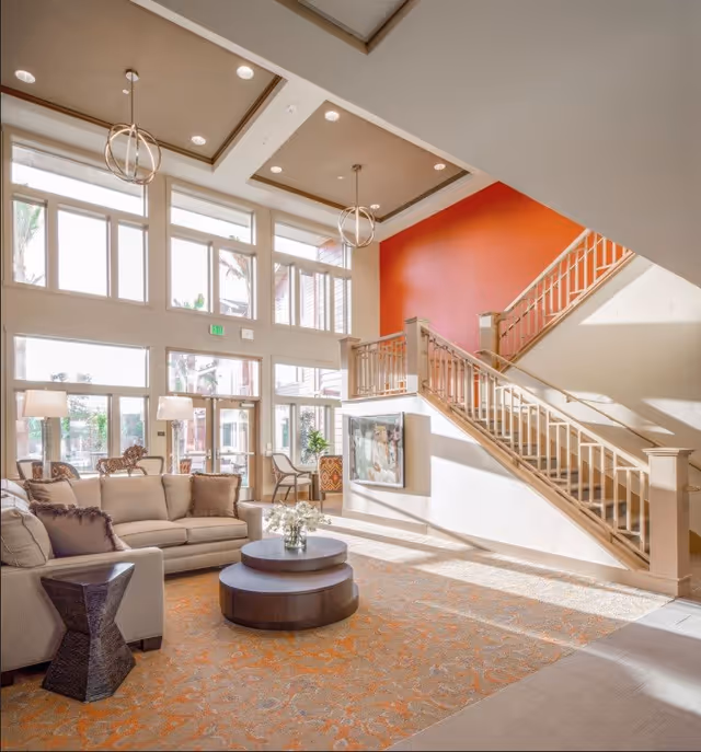 Bright and spacious living room area with large windows allowing natural light to fill the space. The room features a beige sofa with cushions, a round two-tier coffee table with a flower vase, a small dark side table, and two modern pendant lights hanging from a coffered ceiling. A staircase with beige railings leads to an upper floor, and one wall is painted a vibrant orange. There are also chairs and lamps near the windows.