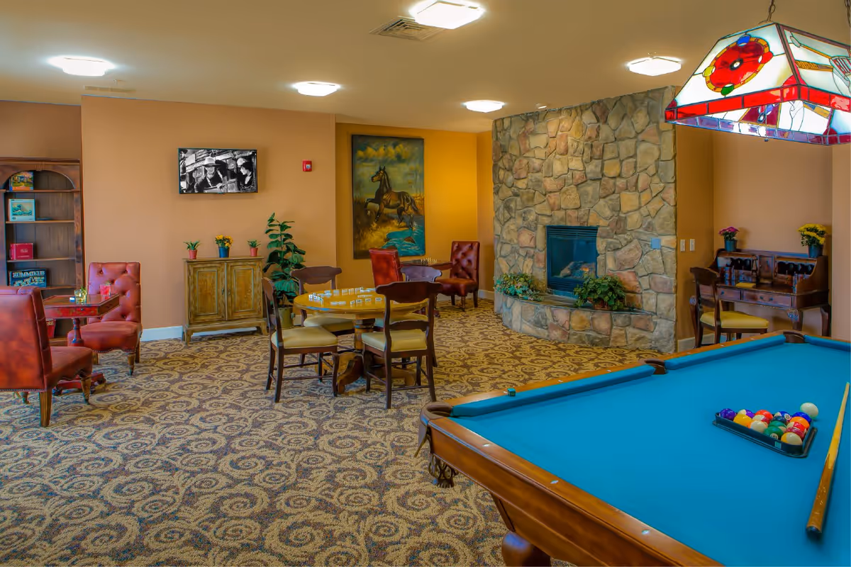 A senior living facility game room featuring a pool table with a rack of billiard balls and a cue stick, a stone fireplace, several tables with chairs including one with a board game, a bookshelf with games, a wall-mounted TV, and decorative plants and artwork on the walls.