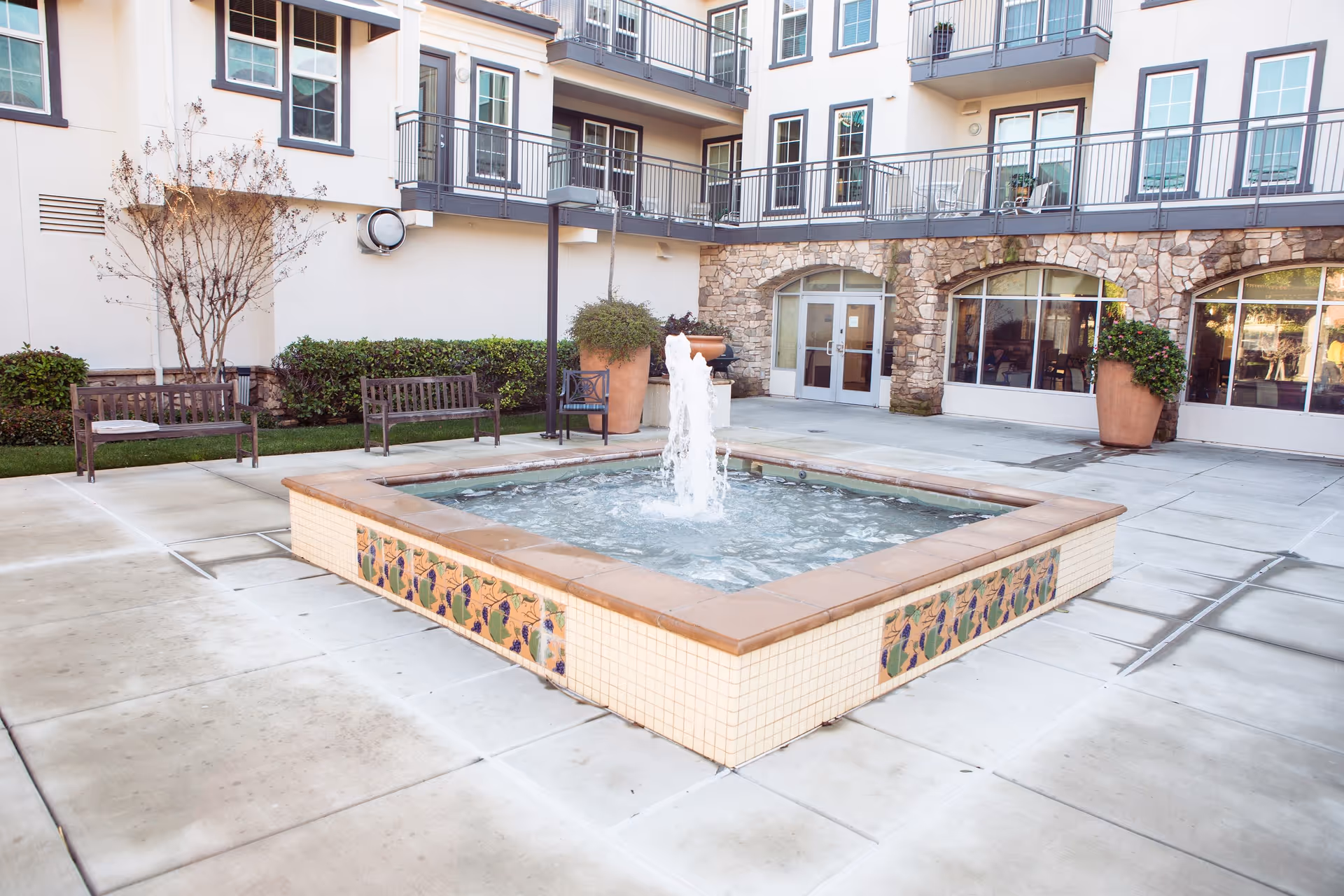 Outdoor courtyard area at Cogir of Brentwood featuring a square water fountain with decorative tiles, surrounded by benches, potted plants, and a multi-story building with balconies and large windows.