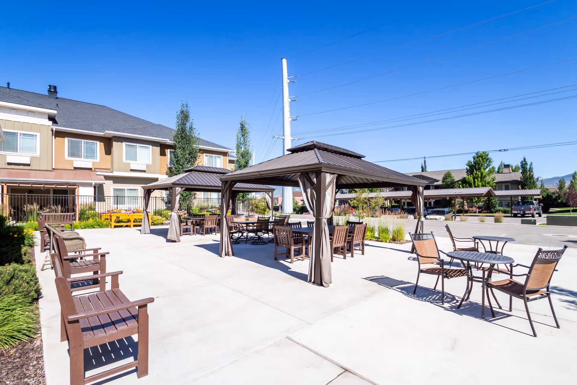 Outdoor patio area at Spring Gardens Senior Living Heber featuring multiple brown wooden benches and metal tables with chairs under two gazebo-style canopies. The area is paved with concrete and surrounded by landscaped greenery and buildings under a clear blue sky.