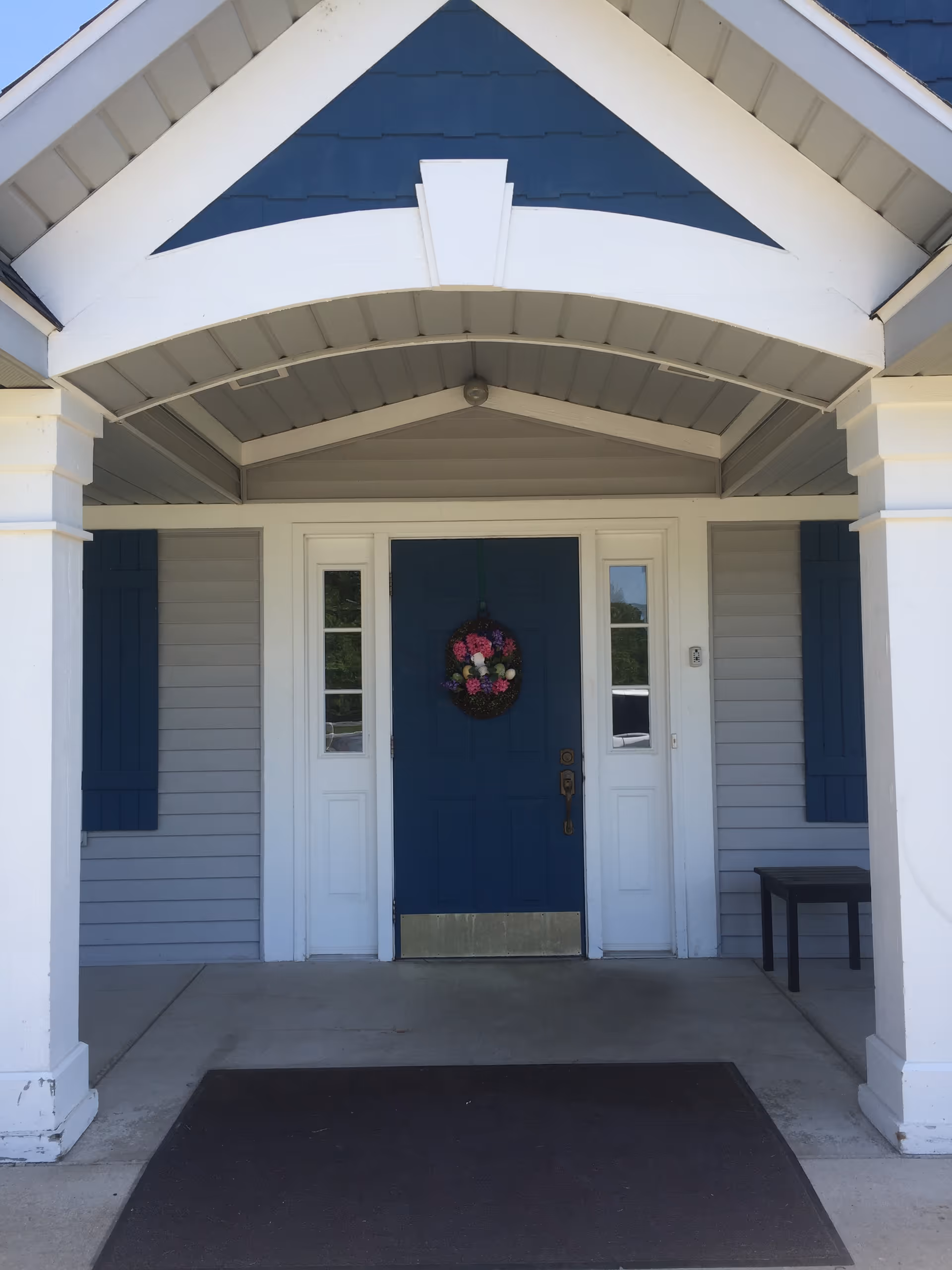 Front entrance of a building with a blue door decorated with a floral wreath, flanked by two narrow windows and blue shutters on a gray exterior wall. The entrance is covered by a white and blue gabled porch with two white columns and a small bench to the right.