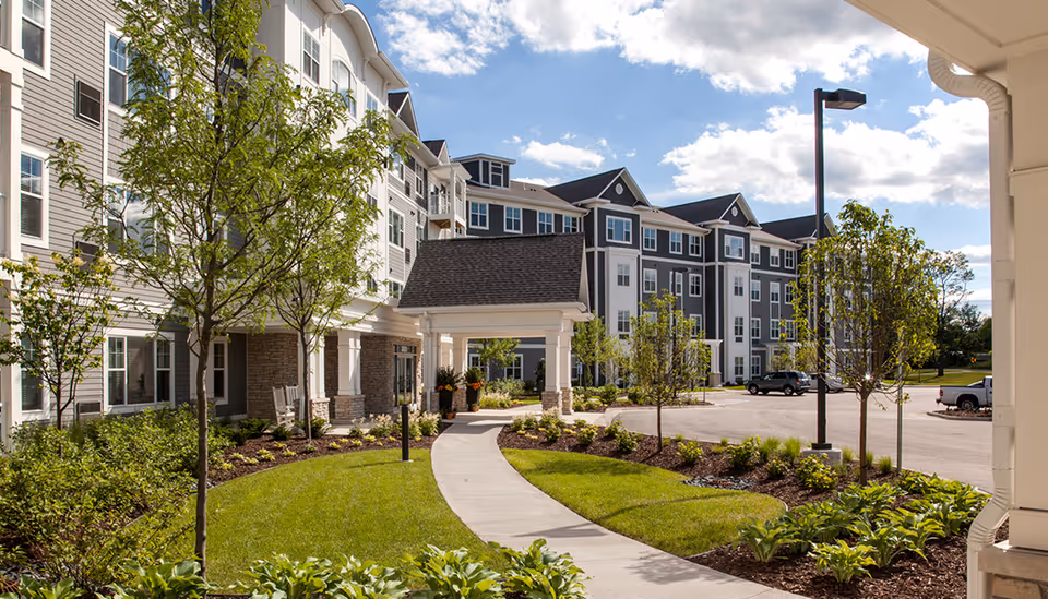 Front exterior of a multi-story senior living building with a covered entrance, walkway, parking area, and landscaped grounds.