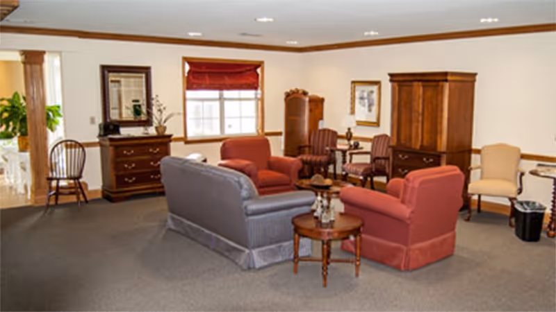 A cozy living room area in an assisted living facility with a gray sofa, two red armchairs, wooden side tables, and additional chairs along the walls. The room has beige walls with wooden trim, a window with a red blind, and wooden furniture including a dresser and a cabinet.